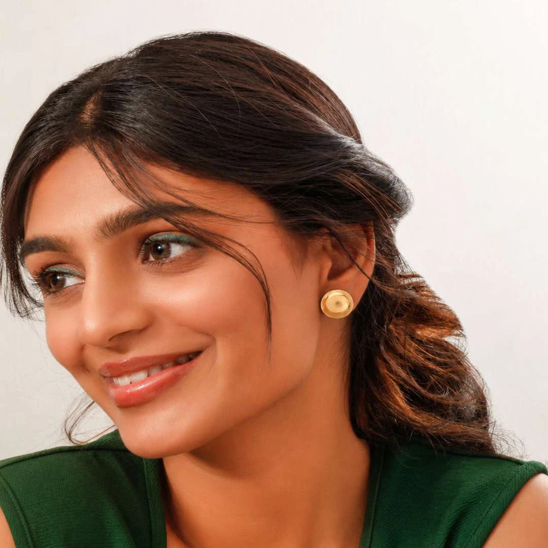 A woman with dark hair pulled back, wearing a green sleeveless top and a small round gold stud earring, shown in a close-up portrait against a plain light background.